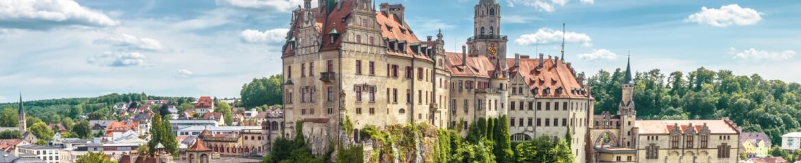 Panoramablick auf ein reich verziertes, historisches Schloss mit roten Ziegeldächern auf einem felsigen Hügel in Baden-Württemberg. Das Schloss überblickt eine charmante Stadt mit farbenfrohen Gebäuden und üppigen grünen Bäumen im Hintergrund unter einem teilweise bewölkten Himmel.