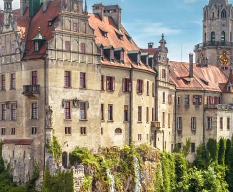 Panoramablick auf ein reich verziertes, historisches Schloss mit roten Ziegeldächern auf einem felsigen Hügel in Baden-Württemberg. Das Schloss überblickt eine charmante Stadt mit farbenfrohen Gebäuden und üppigen grünen Bäumen im Hintergrund unter einem teilweise bewölkten Himmel.