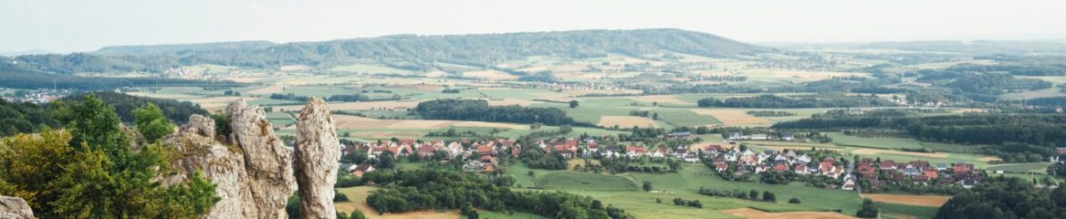 Ein Panoramablick auf eine ländliche Landschaft in Bayern zeigt ein kleines Dorf mit Häusern mit roten Dächern und grünen Feldern, im Hintergrund von sanften Hügeln eingerahmt. Im Vordergrund sind Felsvorsprünge und dichtes Grün zu sehen. Der Himmel ist klar mit einem Hauch von Dunst.