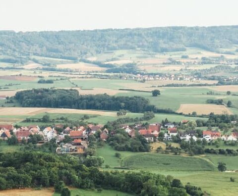 Ein Panoramablick auf eine ländliche Landschaft in Bayern zeigt ein kleines Dorf mit Häusern mit roten Dächern und grünen Feldern, im Hintergrund von sanften Hügeln eingerahmt. Im Vordergrund sind Felsvorsprünge und dichtes Grün zu sehen. Der Himmel ist klar mit einem Hauch von Dunst.