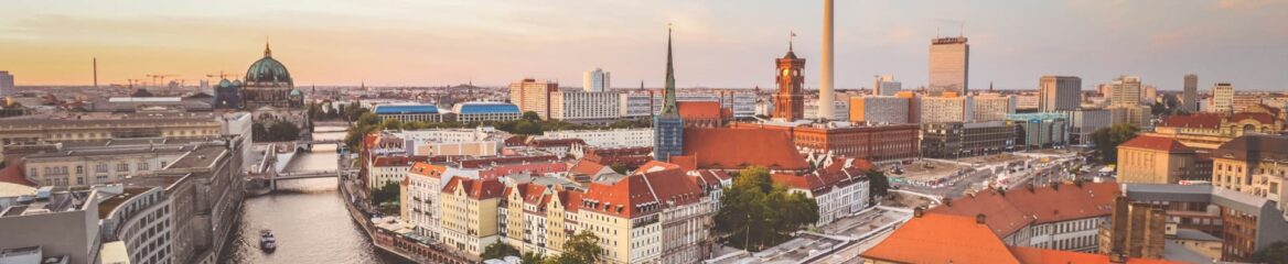 Panoramablick auf die Berliner Stadtlandschaft bei Sonnenuntergang mit historischen und modernen Gebäuden, einem Fluss, der sich durch den Vordergrund schlängelt, und dem berühmten Fernsehturm. Der Himmel ist in warme Orange- und Rosatöne getaucht und wirft einen sanften Glanz über die Stadtlandschaft.