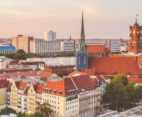 Panoramablick auf die Berliner Stadtlandschaft bei Sonnenuntergang mit historischen und modernen Gebäuden, einem Fluss, der sich durch den Vordergrund schlängelt, und dem berühmten Fernsehturm. Der Himmel ist in warme Orange- und Rosatöne getaucht und wirft einen sanften Glanz über die Stadtlandschaft.