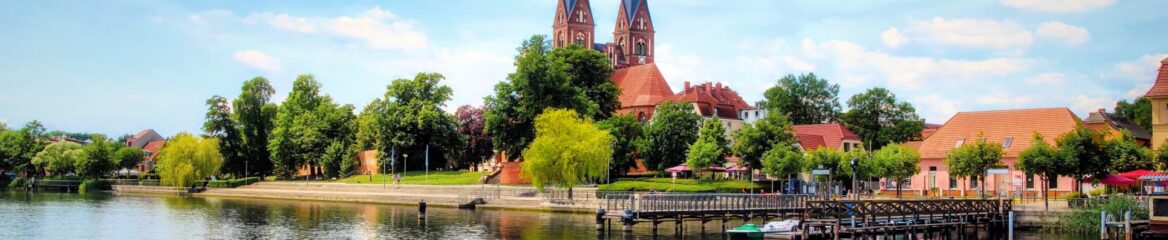 Panoramablick auf eine ruhige Küstenstadt in Brandenburg mit einer von Bäumen gesäumten Promenade, Gebäuden mit roten Dächern und einer markanten Kirche mit zwei Türmen. Die Szene umfasst einen Holzsteg, der sich über ruhiges Wasser unter einem teilweise bewölkten Himmel erstreckt.