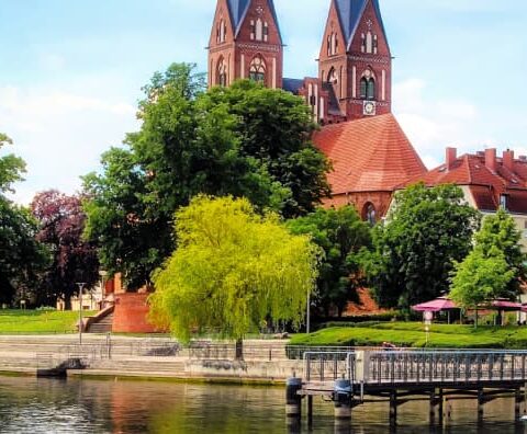 Panoramablick auf eine ruhige Küstenstadt in Brandenburg mit einer von Bäumen gesäumten Promenade, Gebäuden mit roten Dächern und einer markanten Kirche mit zwei Türmen. Die Szene umfasst einen Holzsteg, der sich über ruhiges Wasser unter einem teilweise bewölkten Himmel erstreckt.