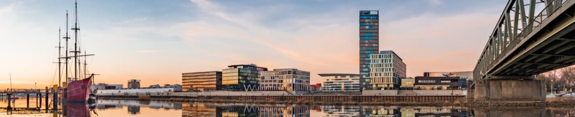 Panoramablick auf die Hafenstadtlandschaft von Bremen bei Sonnenuntergang. Moderne Gebäude mit Glasfassaden, links ein angedocktes Großsegler, ruhiges Wasser, in dem sich die Gebäude spiegeln, und rechts eine grüne Metallbrücke. Der Himmel zeigt Schattierungen von Rosa und Orange.