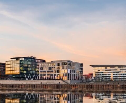 Panoramablick auf die Hafenstadtlandschaft von Bremen bei Sonnenuntergang. Moderne Gebäude mit Glasfassaden, links ein angedocktes Großsegler, ruhiges Wasser, in dem sich die Gebäude spiegeln, und rechts eine grüne Metallbrücke. Der Himmel zeigt Schattierungen von Rosa und Orange.