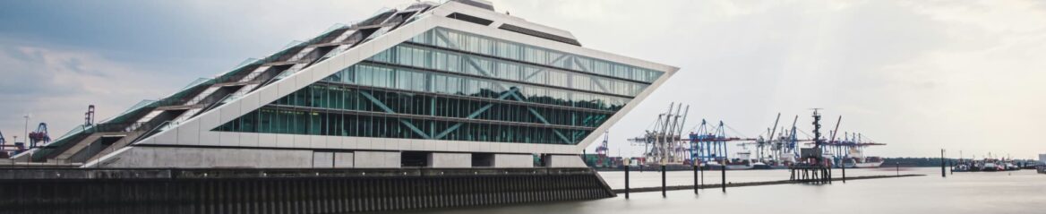 Modernes, eckiges Glasgebäude mit gestufter, pyramidenartiger Struktur neben einem Gewässer in Hamburg. Der Himmel ist bedeckt, im Hintergrund sind angedockte Kräne und Schiffscontainer zu sehen.