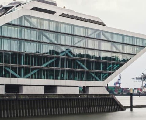 Modernes, eckiges Glasgebäude mit gestufter, pyramidenartiger Struktur neben einem Gewässer in Hamburg. Der Himmel ist bedeckt, im Hintergrund sind angedockte Kräne und Schiffscontainer zu sehen.