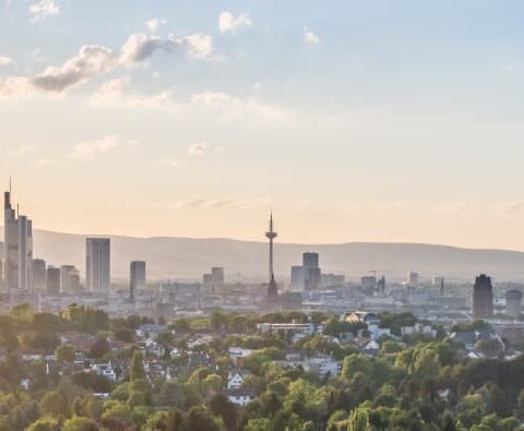 Panoramablick auf die Skyline einer Stadt bei Sonnenuntergang mit zahlreichen modernen Hochhäusern und einem hohen Fernsehturm in Hessen. Im Vordergrund ist eine üppige Grünfläche mit Bäumen zu sehen, während in der Ferne Berge eine malerische Kulisse unter einem Himmel mit vereinzelten Wolken bilden.