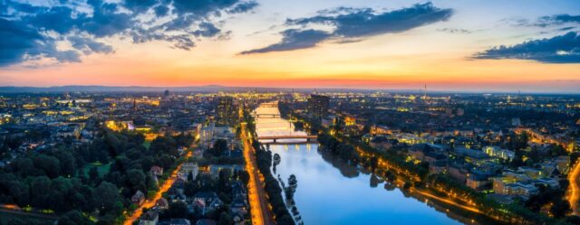 Ein Panoramablick auf eine Stadt in der Abenddämmerung, mit einem gewundenen Fluss, der die Lichter der umliegenden Gebäude und Straßen reflektiert. Der Himmel ist eine Mischung aus Farben der untergehenden Sonne und schafft eine ruhige und doch lebendige Atmosphäre. Bäume und Grünflächen säumen die Flussufer und bieten den Gästen im Monteurzimmer malerische Ausblicke.