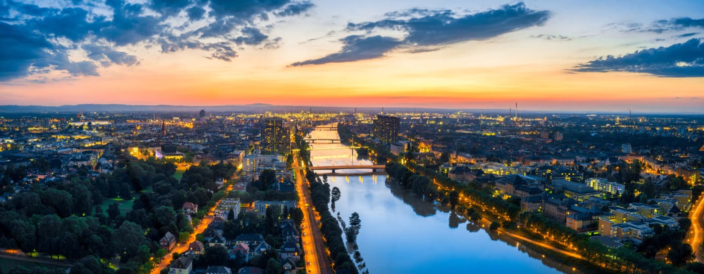 Ein Panoramablick auf eine Stadt in der Abenddämmerung, mit einem gewundenen Fluss, der die Lichter der umliegenden Gebäude und Straßen reflektiert. Der Himmel ist eine Mischung aus Farben der untergehenden Sonne und schafft eine ruhige und doch lebendige Atmosphäre. Bäume und Grünflächen säumen die Flussufer und bieten den Gästen im Monteurzimmer malerische Ausblicke.