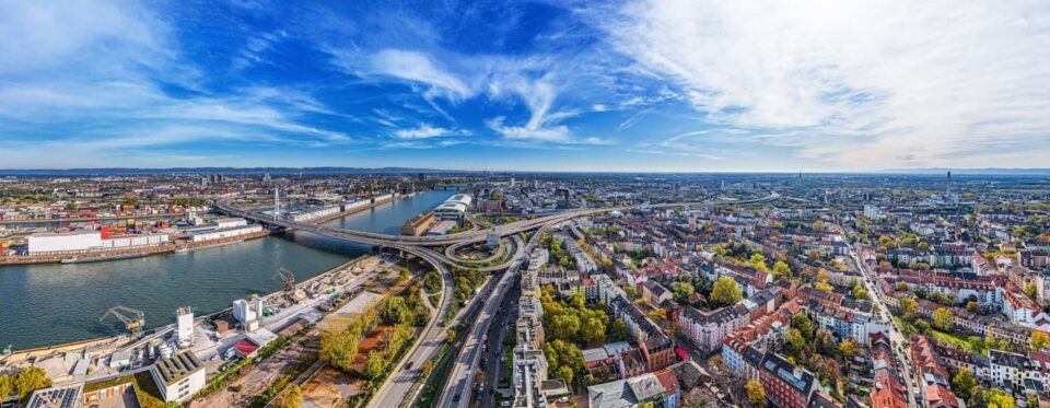 Ein Panoramablick auf eine weitläufige Stadtlandschaft mit einem Fluss, der durch das Zentrum fließt und Brücken freigibt, die verschiedene Teile der Stadt verbinden. Home4Time-Spots wie Handwerkerunterkunft und Monteurwohnung sind zwischen Gebäuden, Grünflächen und Straßen unter einem teilweise bewölkten blauen Himmel mit dünnen Wolken darüber zu sehen.