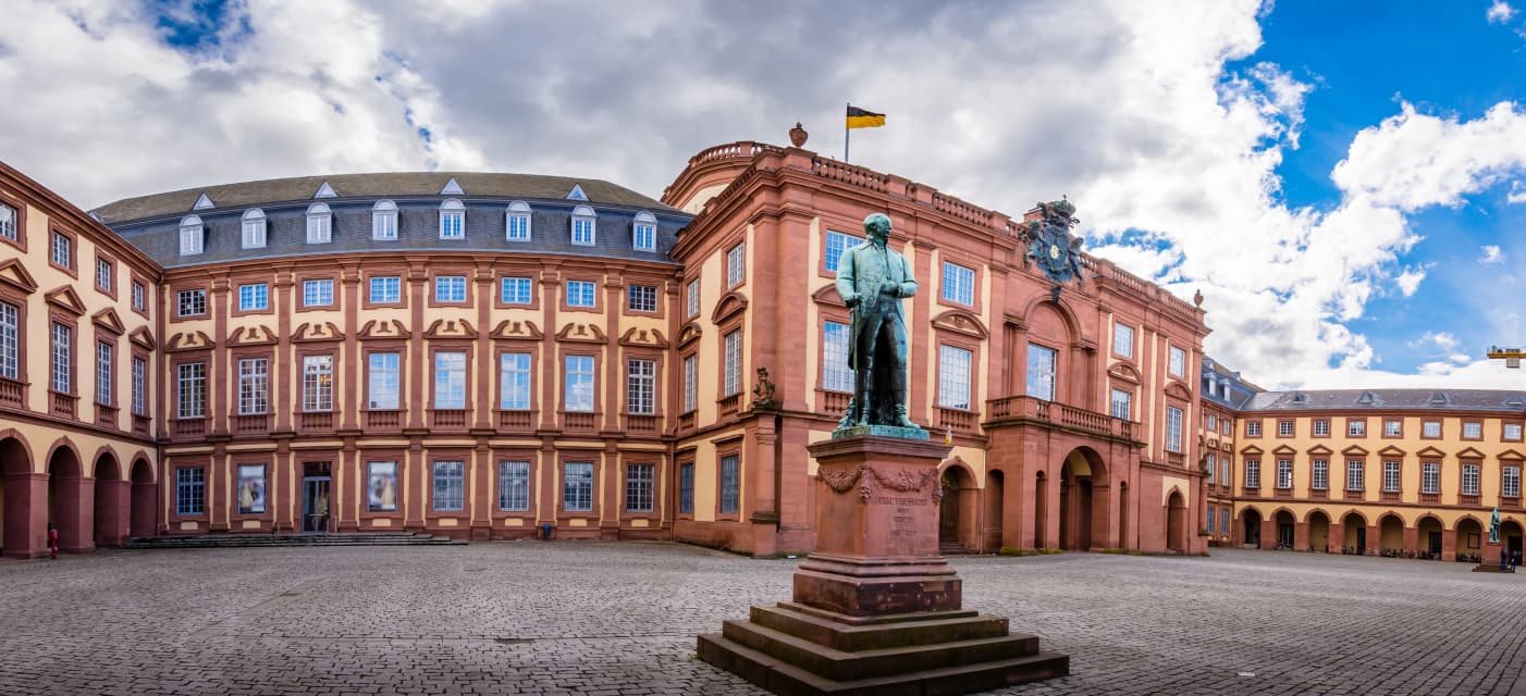 Ein Panoramablick auf den Innenhof des Mannheimer Schlosses in Deutschland zeigt dessen großartige Barockarchitektur, mit einer Statue, die in der Mitte prominent vor einem teilweise bewölkten Himmel prangt.