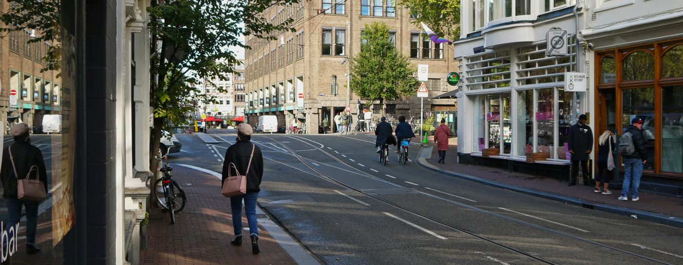 Eine Straßenszene in der Stadt mit Menschen, die zu Fuß gehen und Rad fahren. Die Straße ist gesäumt von Geschäften und Gebäuden, darunter eine Handwerkerunterkunft mit der Aufschrift „Home4Time“, in der ein Geschäft mit einem großen Glasfenster zu sehen ist. Ein paar Bäume sind zu sehen, der Himmel ist klar, und links stehen abgestellte Fahrräder, während in der Mitte Straßenbahnschienen verlaufen.