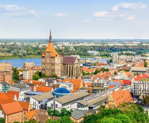 Ein Panoramablick auf eine europäische Stadt, durch die ein Fluss fließt. Das Stadtbild zeichnet sich durch zahlreiche Gebäude mit roten Dächern, Kirchtürme und Grünflächen aus. Im Hintergrund erinnern ausgedehnte Felder und ein klarer blauer Himmel mit einigen Wolken an den Charme Mecklenburg-Vorpommerns.