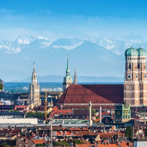 Panoramablick auf München, Deutschland, mit der Frauenkirche mit ihren markanten Zwillingskuppeln, historischen Gebäuden und den bayerischen Alpen im Hintergrund unter einem klaren blauen Himmel. Die Skyline von München verbindet architektonische Wahrzeichen mit schneebedeckten Bergen in der Ferne.