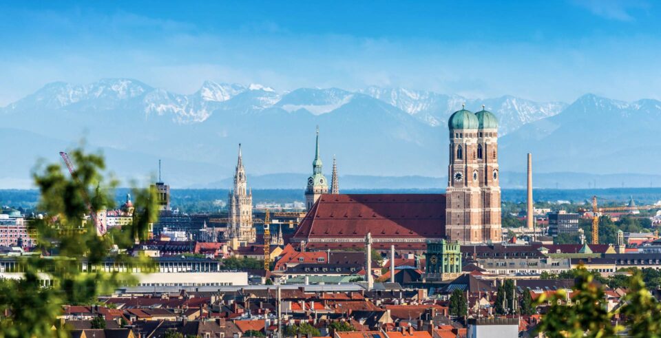 Panoramablick auf München, Deutschland, mit der Frauenkirche mit ihren markanten Zwillingskuppeln, historischen Gebäuden und den bayerischen Alpen im Hintergrund unter einem klaren blauen Himmel. Die Skyline von München verbindet architektonische Wahrzeichen mit schneebedeckten Bergen in der Ferne.