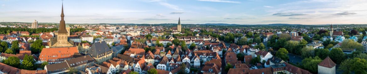 Eine Panorama-Luftaufnahme einer europäischen Stadtlandschaft mit mehreren Kirchtürmen, Gebäuden mit roten Dächern und von Bäumen gesäumten Straßen unter einem blauen Himmel mit vereinzelten Wolken. Im Hintergrund sind sanfte Hügel zu sehen.