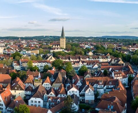 Eine Panorama-Luftaufnahme einer europäischen Stadtlandschaft mit mehreren Kirchtürmen, Gebäuden mit roten Dächern und von Bäumen gesäumten Straßen unter einem blauen Himmel mit vereinzelten Wolken. Im Hintergrund sind sanfte Hügel zu sehen.