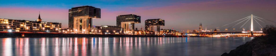 Panoramablick auf die moderne Uferpromenade von Köln in der Abenddämmerung mit beleuchteten Gebäuden, darunter die markanten Kranenhäuser. Die Schrägseilbrücke und der Kölner Dom in der Ferne ergänzen die atemberaubende Skyline von Nordrhein-Westfalen und spiegeln sich im ruhigen Rhein darunter wider.