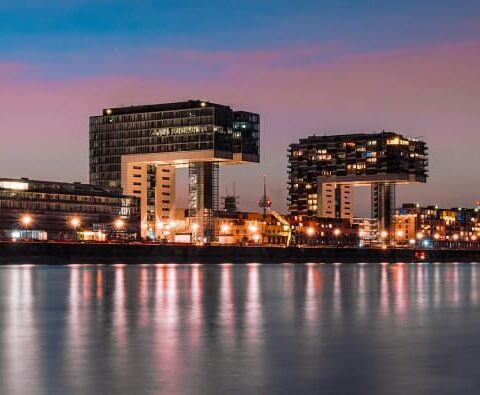 Panoramablick auf die moderne Uferpromenade von Köln in der Abenddämmerung mit beleuchteten Gebäuden, darunter die markanten Kranenhäuser. Die Schrägseilbrücke und der Kölner Dom in der Ferne ergänzen die atemberaubende Skyline von Nordrhein-Westfalen und spiegeln sich im ruhigen Rhein darunter wider.