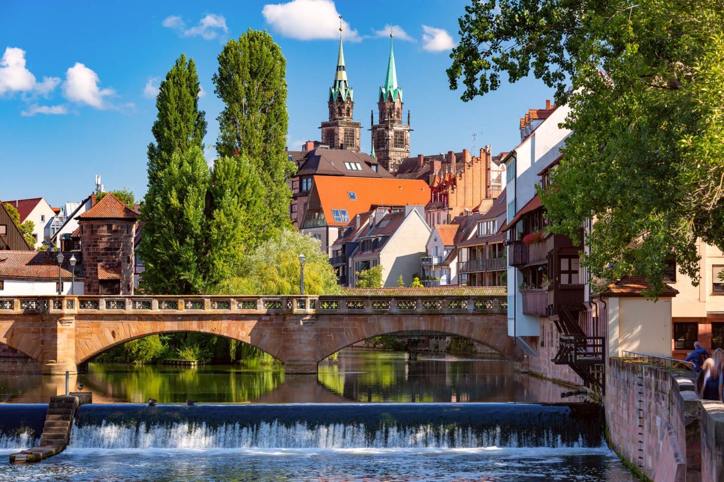 Eine malerische Szene aus Nürnberg, Deutschland, mit einer historischen Steinbrücke über einem fließenden Fluss mit einem kleinen Wasserfall. Im Hintergrund sind charmante alte Gebäude mit spitzen Dächern und Kirchtürmen zu sehen, umgeben von üppigen grünen Bäumen unter einem strahlend blauen Himmel.