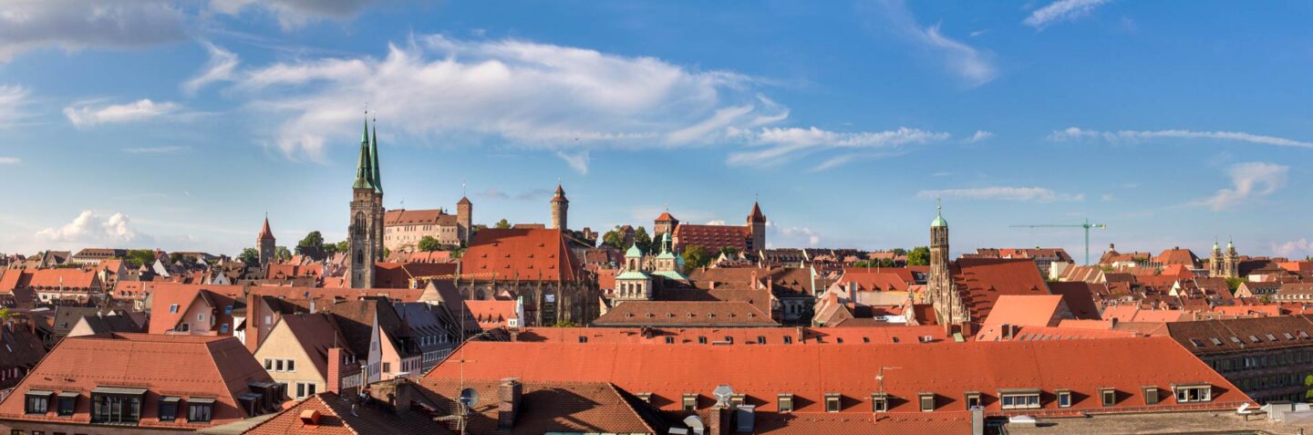 Panoramablick auf die Nürnberger Stadtlandschaft mit zahlreichen Gebäuden mit roten Dächern unter einem blauen Himmel mit vereinzelten Wolken. Zu den markanten Bauwerken gehören eine hohe Kirche mit Turm und mehrere historische Gebäude im Hintergrund. In der Ferne sind Bäume und Grünflächen zu sehen.