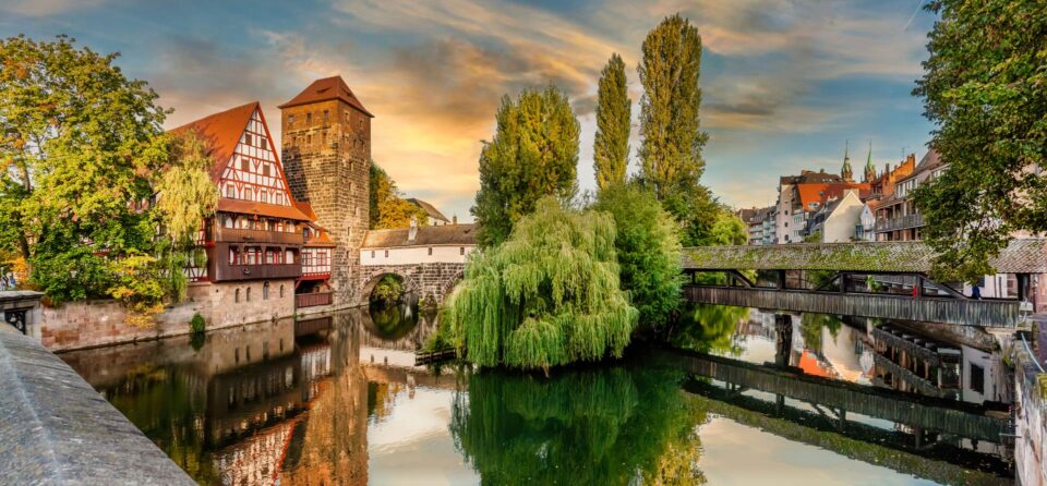 Ein malerischer Blick auf das historische Nürnberg bei Sonnenuntergang zeigt Fachwerkhäuser, einen Steinturm und üppiges Grün, das sich in einem ruhigen Fluss spiegelt. Eine Brücke überquert den Fluss und verbindet Teile der Stadt. Bäume und Gebäude vervollständigen die ruhige Szene.