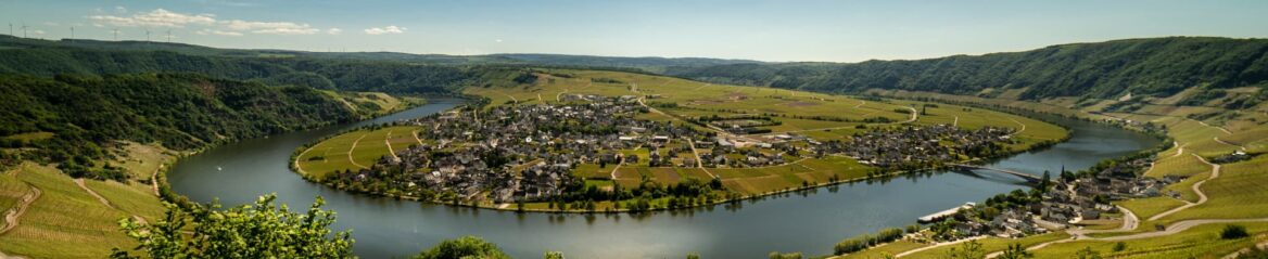 Panoramablick auf ein malerisches Dorf am Flussufer, eingebettet in die üppigen, sanften Hügel von Rheinland-Pfalz. Die ruhige Mosel schlängelt sich um die Siedlung und schafft eine idyllische Szenerie aus Grün und Ruhe, wobei eine Brücke beide Seiten der Landschaft verbindet.