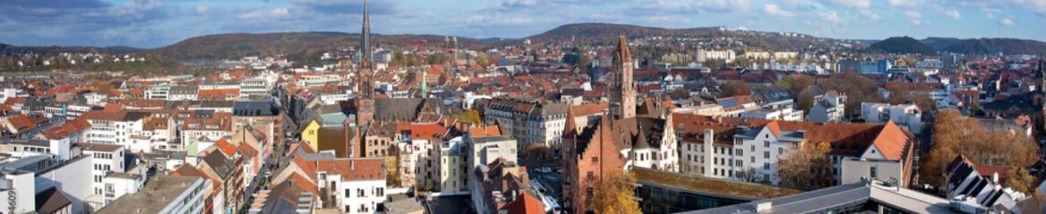 Ein Panoramablick auf eine europäische Stadt im Saarland zeigt historische Gebäude mit roten Ziegeldächern, eine zentrale Kirche mit einem hohen Turm und umliegende Hügel unter einem teilweise bewölkten Himmel.