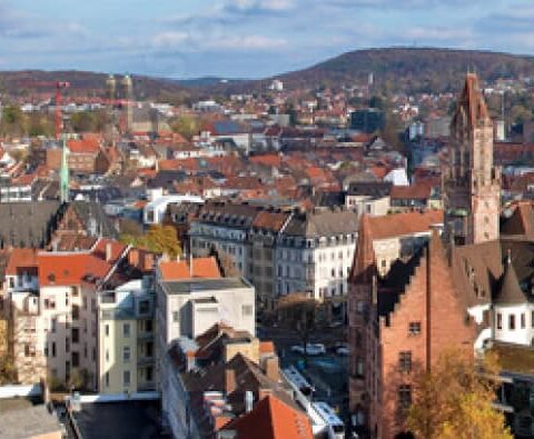 Ein Panoramablick auf eine europäische Stadt im Saarland zeigt historische Gebäude mit roten Ziegeldächern, eine zentrale Kirche mit einem hohen Turm und umliegende Hügel unter einem teilweise bewölkten Himmel.