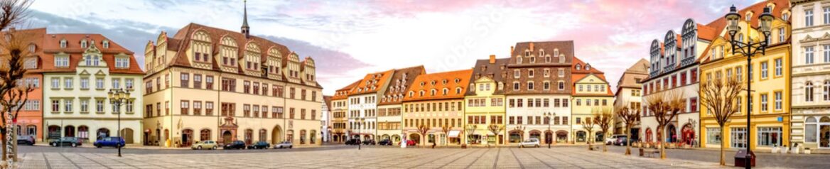 Panoramablick auf einen malerischen europäischen Stadtplatz in Sachsen-Anhalt, umgeben von farbenfrohen, historischen Gebäuden mit kunstvollen architektonischen Details. Auf dem Platz parken Autos und es gibt einige Fußgänger, im Hintergrund ein rosafarbener Abendhimmel.