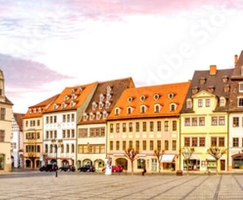 Panoramablick auf einen malerischen europäischen Stadtplatz in Sachsen-Anhalt, umgeben von farbenfrohen, historischen Gebäuden mit kunstvollen architektonischen Details. Auf dem Platz parken Autos und es gibt einige Fußgänger, im Hintergrund ein rosafarbener Abendhimmel.