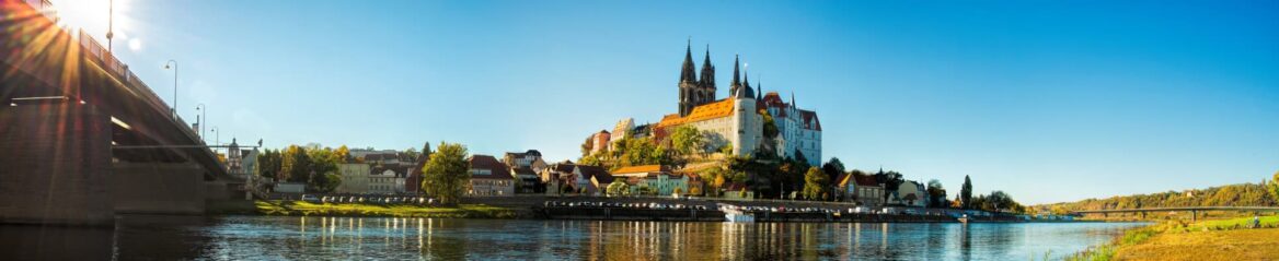 Panoramablick auf die Albrechtsburg und den Meißner Dom in Meißen, Sachsen, Deutschland, mit der Elbe im Vordergrund an einem klaren, sonnigen Tag. Die historischen Gebäude der Stadt säumen das Flussufer und links überspannt eine Brücke das Wasser.