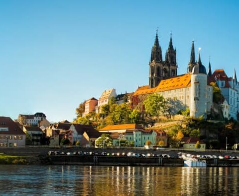 Panoramablick auf die Albrechtsburg und den Meißner Dom in Meißen, Sachsen, Deutschland, mit der Elbe im Vordergrund an einem klaren, sonnigen Tag. Die historischen Gebäude der Stadt säumen das Flussufer und links überspannt eine Brücke das Wasser.