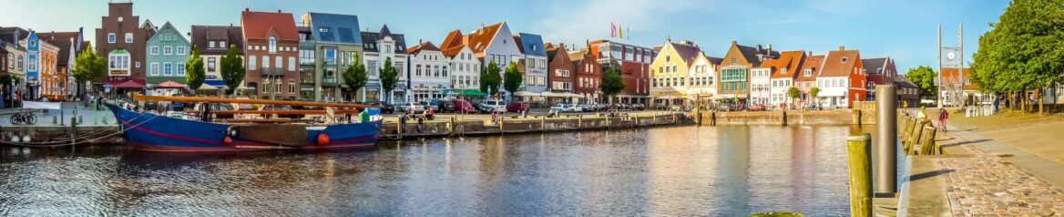 Ein Panoramablick auf eine malerische Küstenstadt in Schleswig-Holstein mit farbenfrohen historischen Gebäuden entlang der ruhigen Wasserstraße. Ein großes Holzboot liegt vor Anker und Menschen schlendern am Kai entlang. Der klare blaue Himmel verstärkt die lebendigen Farben und die bezaubernde Atmosphäre der Szene.