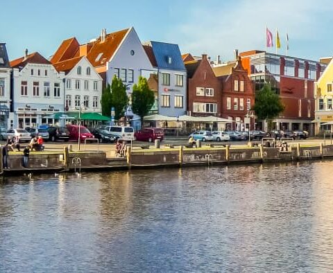 Ein Panoramablick auf eine malerische Küstenstadt in Schleswig-Holstein mit farbenfrohen historischen Gebäuden entlang der ruhigen Wasserstraße. Ein großes Holzboot liegt vor Anker und Menschen schlendern am Kai entlang. Der klare blaue Himmel verstärkt die lebendigen Farben und die bezaubernde Atmosphäre der Szene.