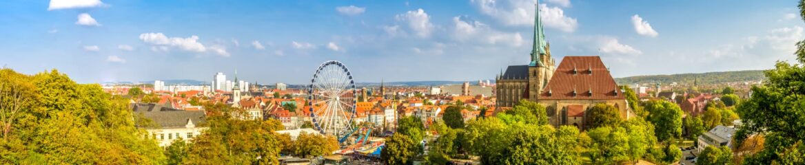 Panoramablick auf eine Stadtlandschaft mit einem Riesenrad, historischen Gebäuden und einer großen Kirche mit grünen Türmen. Die Szene ist von üppigem Grün unter einem blauen Himmel mit vereinzelten Wolken umgeben. Im Hintergrund sind moderne Gebäude und entfernte Hügel sichtbar.