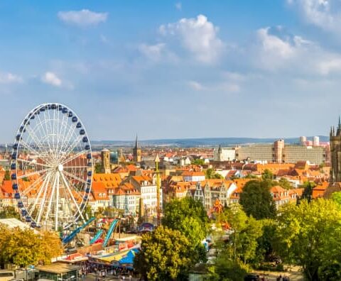 Panoramablick auf eine Stadtlandschaft mit einem Riesenrad, historischen Gebäuden und einer großen Kirche mit grünen Türmen. Die Szene ist von üppigem Grün unter einem blauen Himmel mit vereinzelten Wolken umgeben. Im Hintergrund sind moderne Gebäude und entfernte Hügel sichtbar.