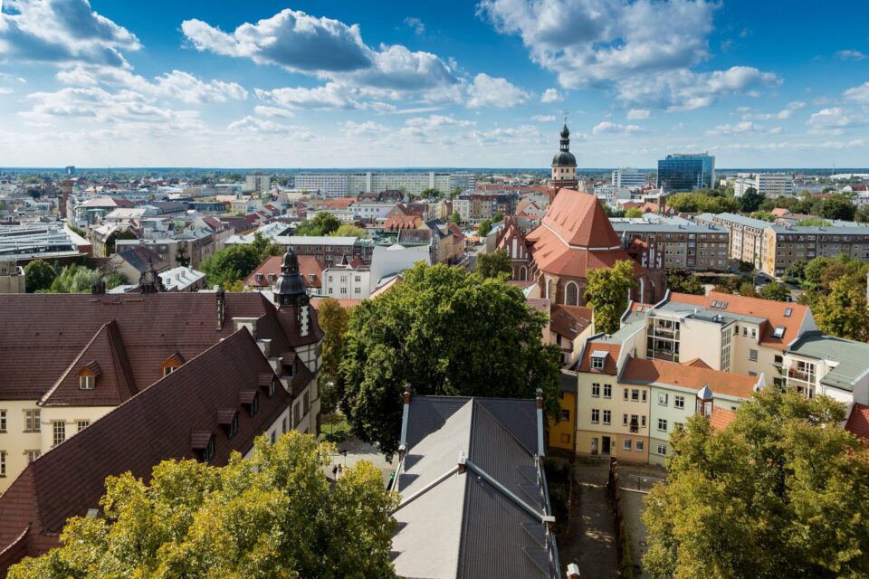 Ein Blick von oben auf die Stadt zeigt eine Mischung aus modernen und traditionellen Gebäuden unter einem klaren blauen Himmel mit vereinzelten Wolken. Inmitten dieses Cottbus-Panoramas sind zwischen den Gebäuden eine markante Kirche mit rotem Dach und verschiedene grüne Bäume zu sehen.