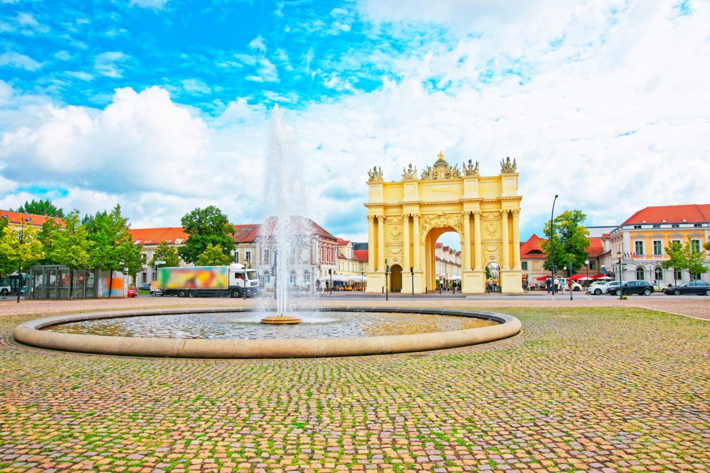 Ein großer Brunnen in einem runden Becken steht zentral auf einem Kopfsteinpflasterplatz in Potsdam. Dahinter befindet sich ein verzierter, gelber Triumphbogen. Im Hintergrund sind historische Gebäude mit roten Dächern und ein teilweise bewölkter blauer Himmel zu sehen. Fahrzeuge und Bäume sind rundherum zu sehen, was den Platz perfekt für Reisebegeisterte und Tourismusfotografie macht.