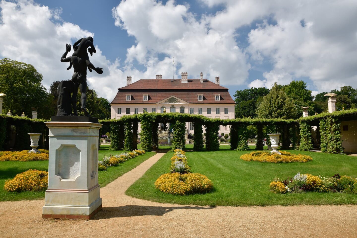 Vor einem großen, stattlichen Gebäude mit rotem Dach und mehreren Schornsteinen, das an die prachtvolle Architektur von Cottbus erinnert, liegt ein kunstvoller Garten mit gepflegten Rasenflächen und Blumenbeeten. Im Vordergrund steht eine schwarze Statue auf einem Sockel, umgeben von üppigen grünen Bäumen und Hecken.