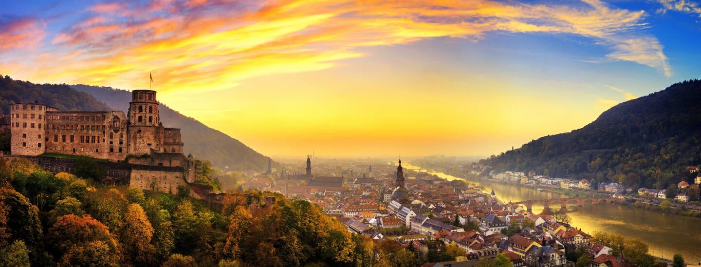 Panoramablick auf Heidelberg bei Sonnenuntergang. Das Bild zeigt links das Heidelberger Schloss mit der historischen Innenstadt und der Alten Brücke über den Neckar. Der Himmel ist in leuchtenden Orange-, Gelb- und Blautönen gehalten und verleiht dieser bezaubernden Heidelberger Szene Wärme.