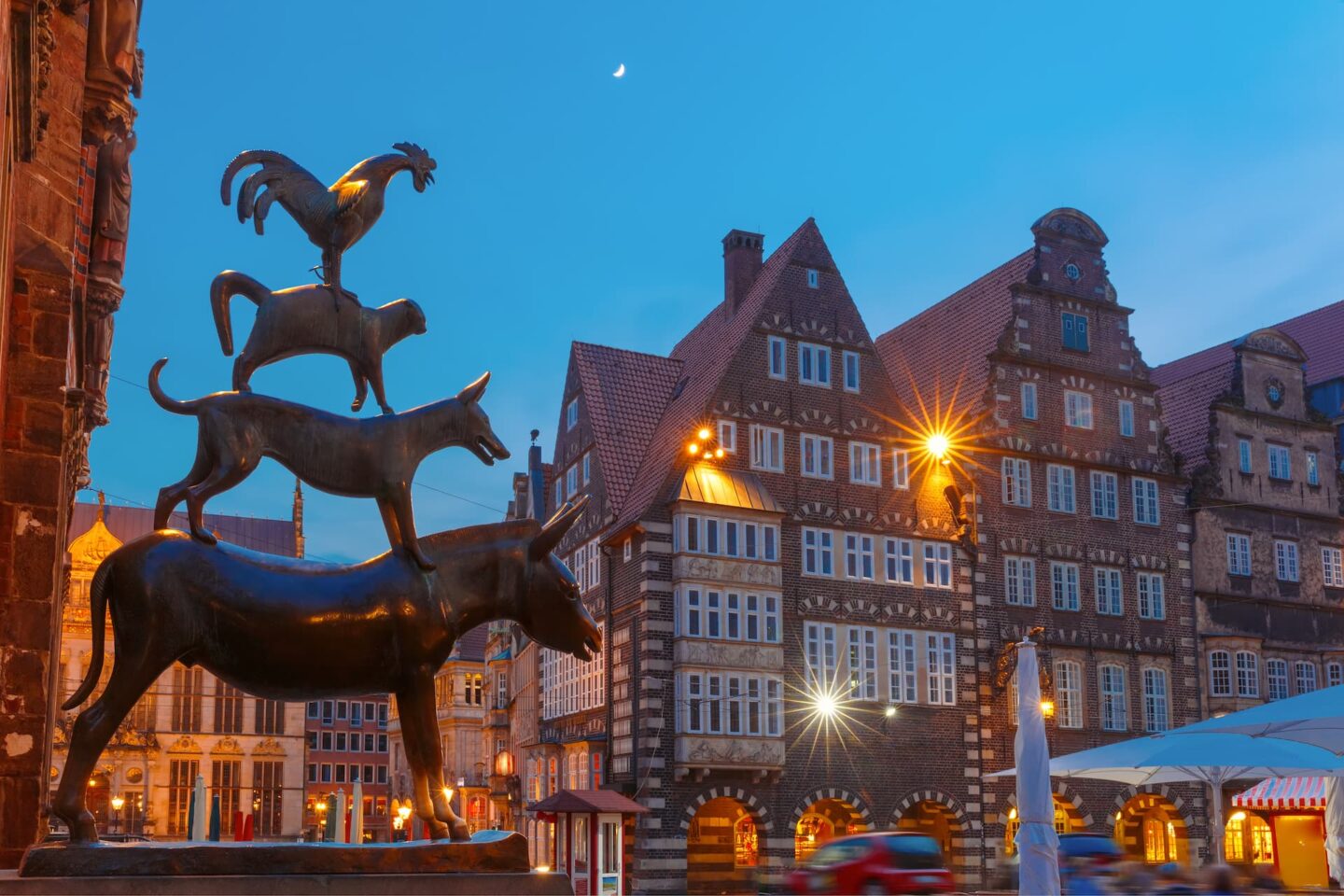 Das Bild zeigt die Bronzestatue der Bremer Stadtmusikanten – vier übereinander gestapelte Tiere – in Bremen, Deutschland. Im Hintergrund sind historische, beleuchtete Gebäude des Bremer Marktplatzes in der Abenddämmerung mit einer Mondsichel am Himmel zu sehen.