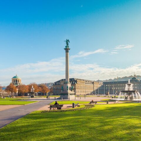 Ein weiter Blick auf den Schlossplatz in Stuttgart an einem sonnigen Tag. Das Bild zeigt ausgedehnte grüne Rasenflächen, die Jubiläumssäule mit der Concordia-Statue, Brunnen, Bänke und historische Gebäude rund um den Platz. Der klare blaue Himmel verstärkt Stuttgarts architektonischen Charme.