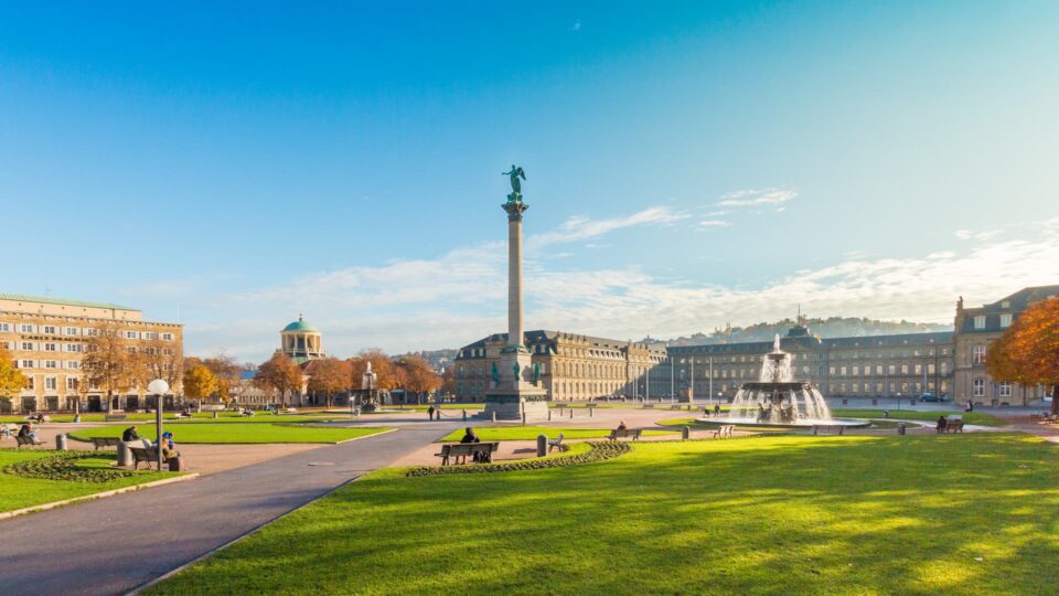 Ein weiter Blick auf den Schlossplatz in Stuttgart an einem sonnigen Tag. Das Bild zeigt ausgedehnte grüne Rasenflächen, die Jubiläumssäule mit der Concordia-Statue, Brunnen, Bänke und historische Gebäude rund um den Platz. Der klare blaue Himmel verstärkt Stuttgarts architektonischen Charme.