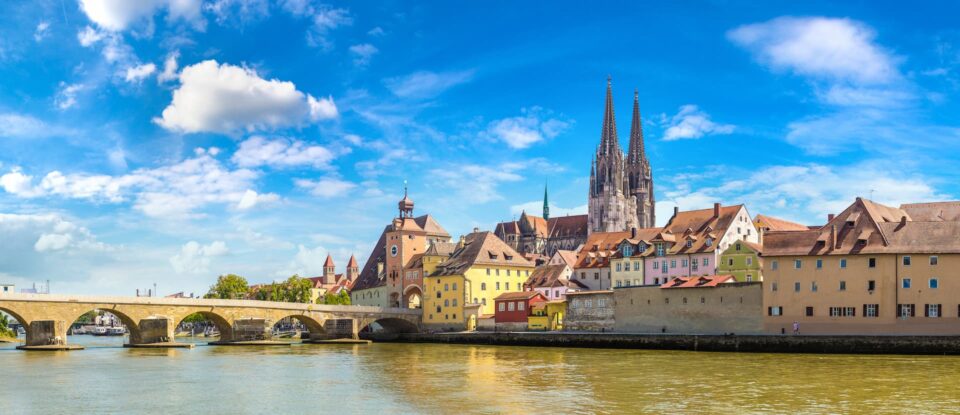 Panoramablick auf eine malerische mittelalterliche Stadt in Bayern mit farbenfrohen Gebäuden entlang eines Flusses. Eine Steinbrücke überspannt den Fluss und ein Dom mit hohen Türmen dominiert die Skyline von Regensburg unter einem strahlend blauen Himmel mit vereinzelten Wolken, was es zu einem idealen Ort für den Tourismus macht.