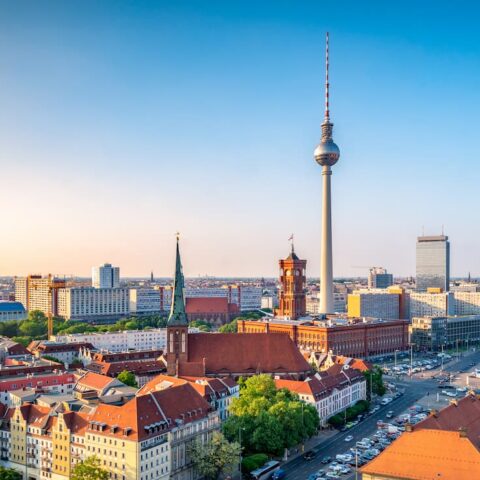 Panoramablick auf Berlin bei Sonnenaufgang, perfekt für Reiseliebhaber, mit dem ikonischen Fernsehturm in der Mitte. Historische Gebäude, moderne Wolkenkratzer und ein gewundener Fluss sind zu sehen, Vögel fliegen über einen klaren Himmel. Die Stadt ist in sanftes, frühes Morgenlicht getaucht.