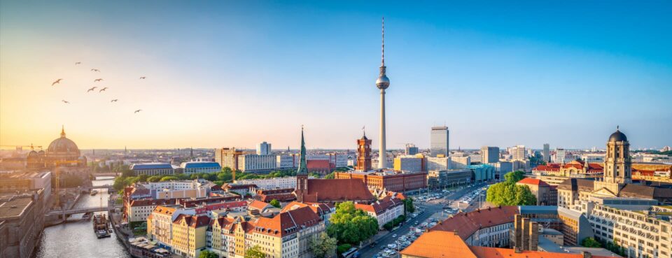 Panoramablick auf Berlin bei Sonnenaufgang, perfekt für Reiseliebhaber, mit dem ikonischen Fernsehturm in der Mitte. Historische Gebäude, moderne Wolkenkratzer und ein gewundener Fluss sind zu sehen, Vögel fliegen über einen klaren Himmel. Die Stadt ist in sanftes, frühes Morgenlicht getaucht.