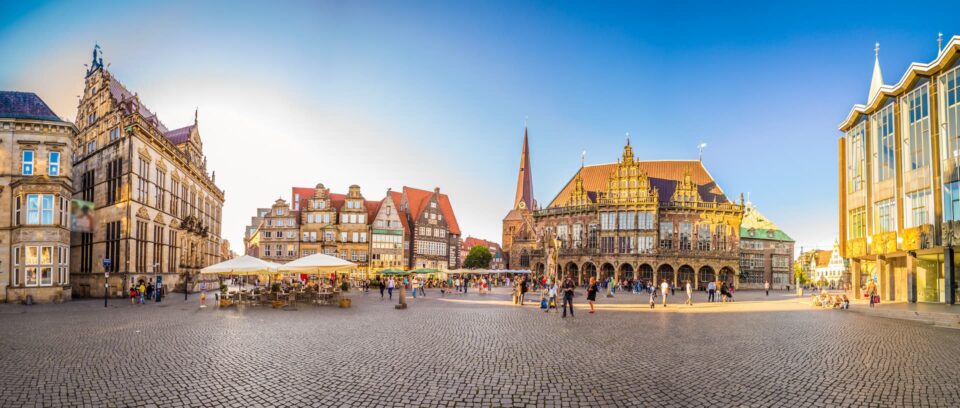 Panoramablick auf den historischen Stadtplatz von Bremen mit Kopfsteinpflaster, Straßencafés und flanierenden Menschen. Zu den umliegenden Gebäuden gehören eine gotische Kirche mit einem hohen Turm und verschiedene kunstvolle Gebäude mit roten Ziegeldächern und kunstvollen Fassaden vor einem klaren blauen Himmel.